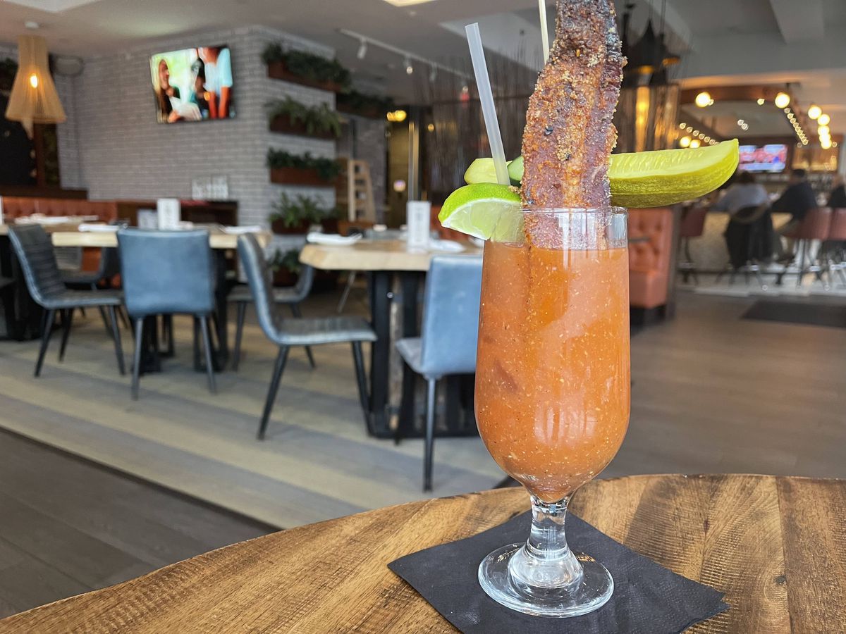 A glass filled with a red cocktail and garnished with a slice of bacon sits on a wooden table.