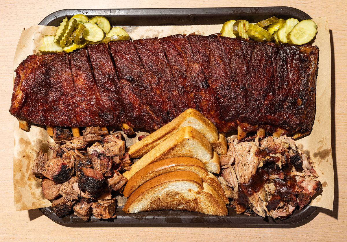 From above, a tray of barbecued meat, including a full rack of ribs, burnt ends, and pulled pork, along with slices of white bread toast. 