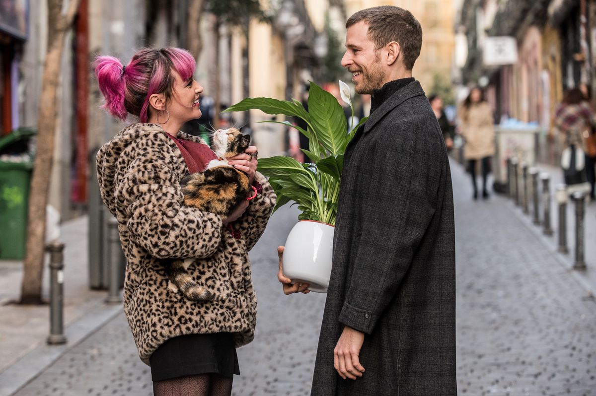 A couple stands together on the street in Love at First Kiss. On the left, a person with pink hair wears a leopard print coat and holds a cat. On the right, a person wears a long black coat and holds a potted plant