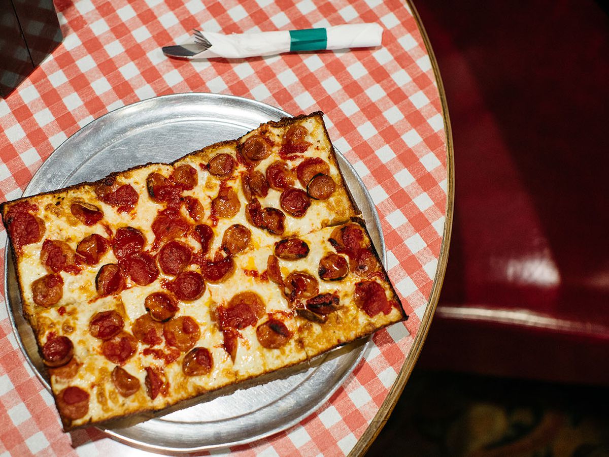 A square pizza with pepperoni on a round table with a red and white checked tablecloth.