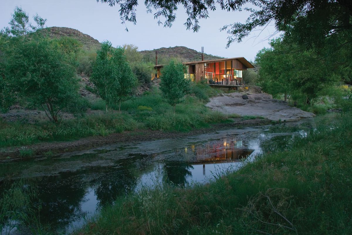Wide shot of modern home with broadly sloping A-frame roof with glass wall overlooking nature and a stream in the foreground.