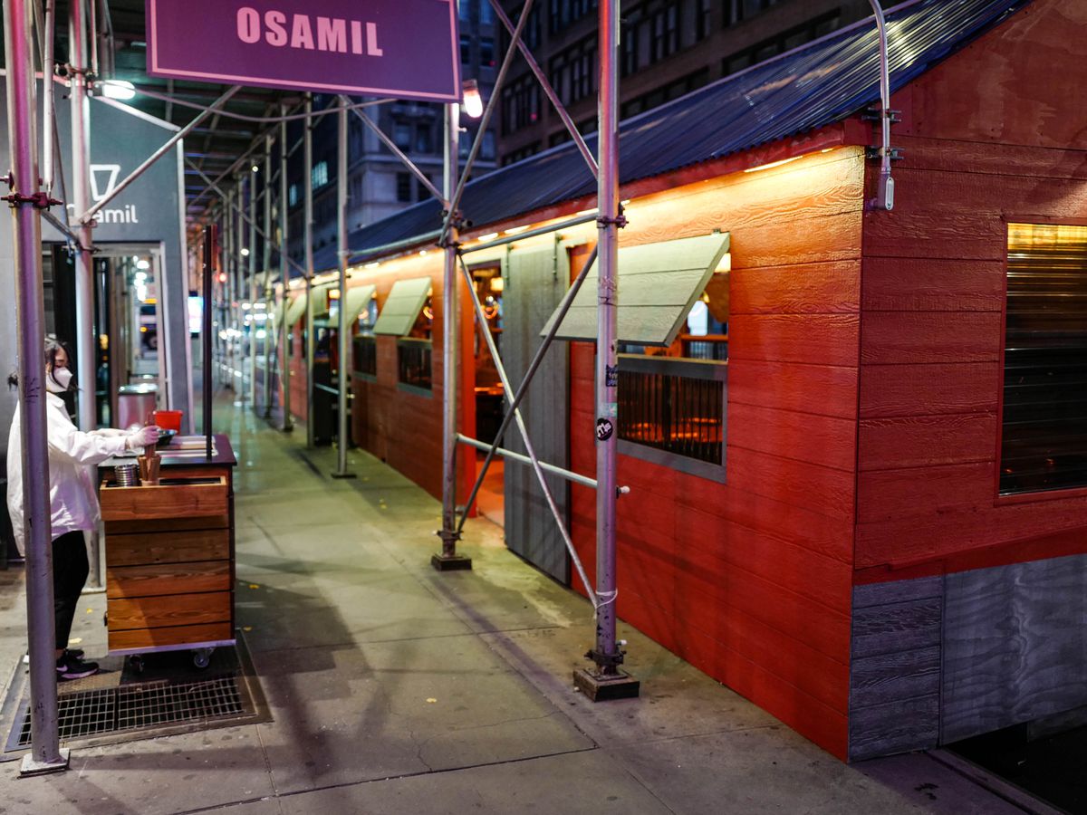 An outdoor dining structure in front of a restaurant is painted red with a blue roof. Beside it, a host stands at a counter under a sign that reads “Osamil.”