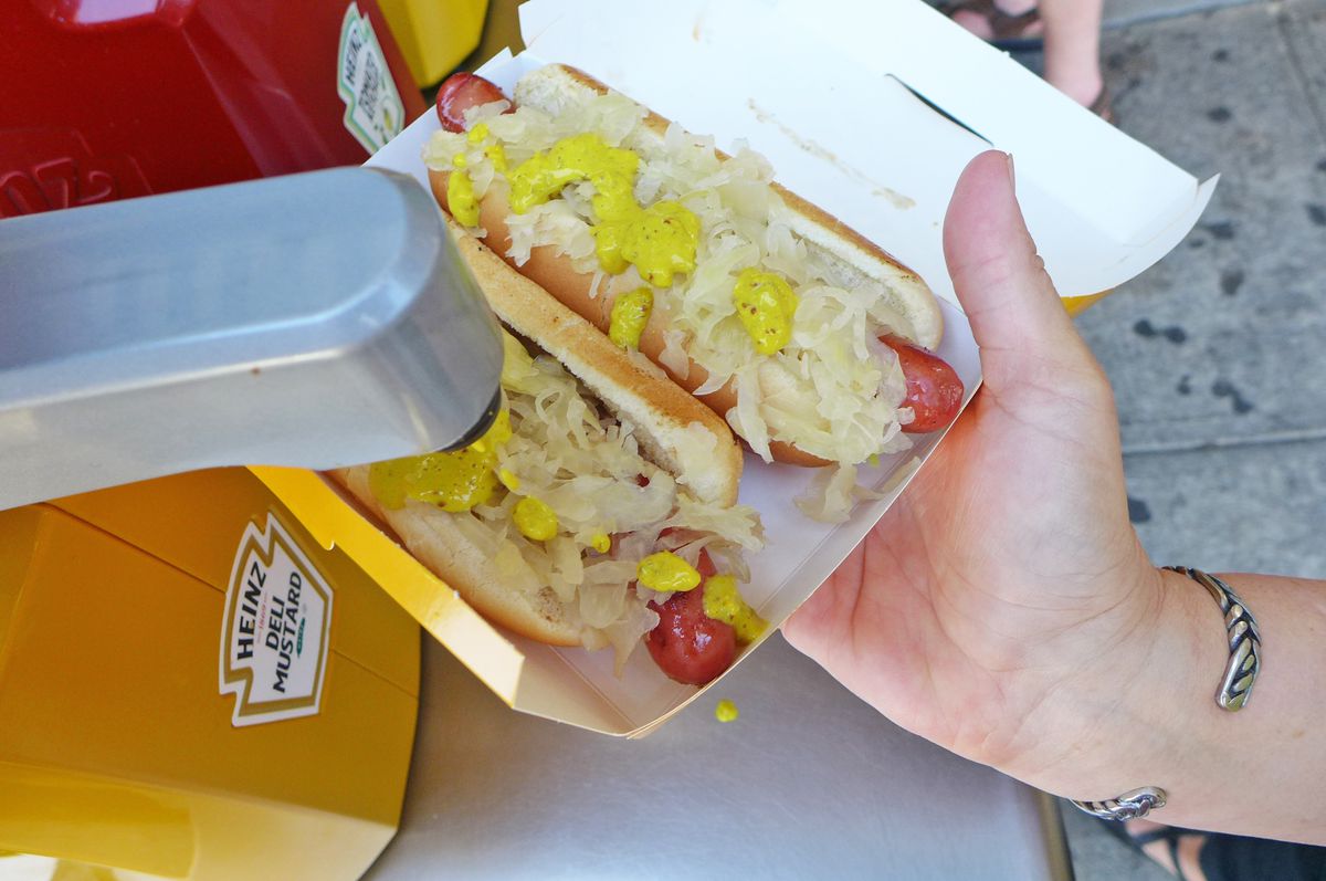 Aa hand holds two hot dogs in buns in paper containers about to apply mustard.