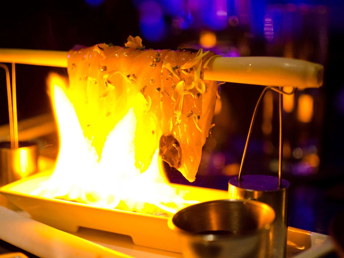 Raw salmon is draped over a wooden dowel suspended above a fire, which is searing the fish