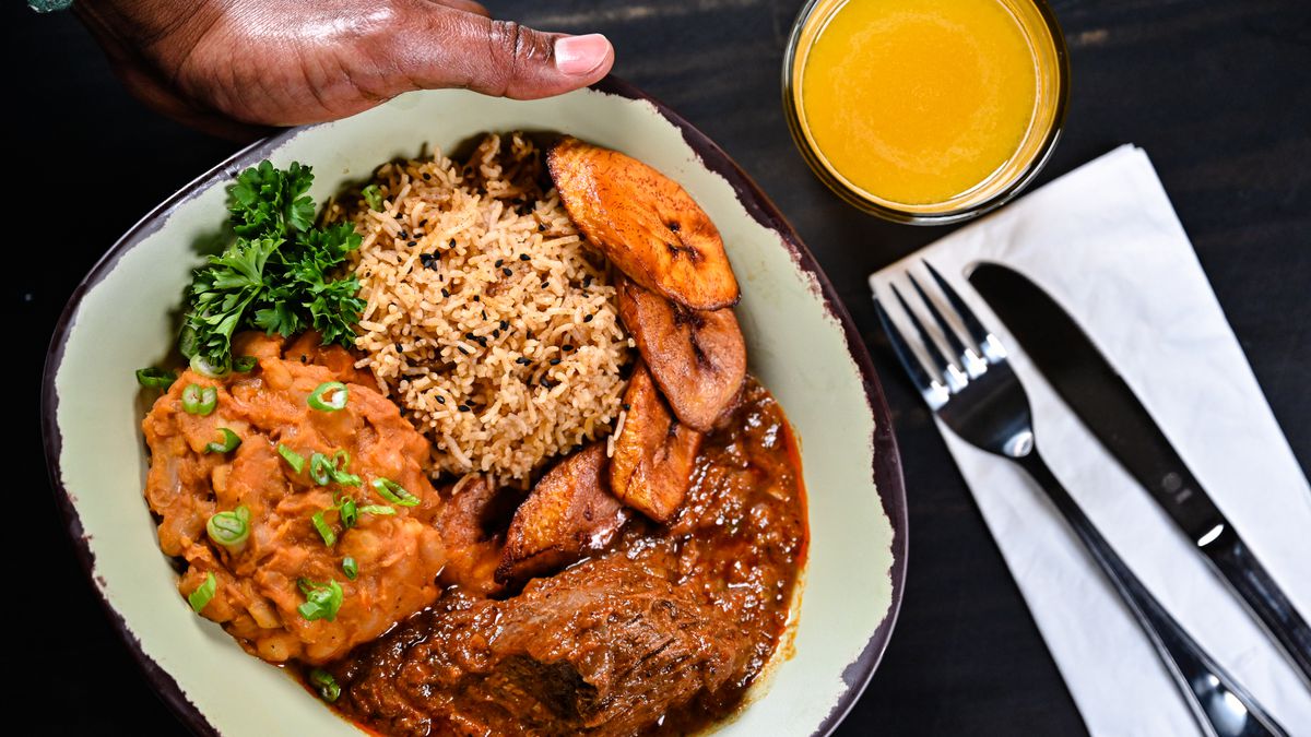 A hand holds a bowl filled with Nyumbani filled with several stews and slice plantains with a glass of yellow fruit juice next to it.