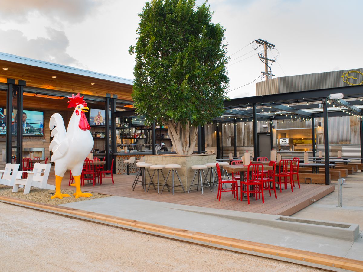 The outdoor dining area of Crack Shack has a giant rooster, tables with red chairs and a large tree with seating around it.
