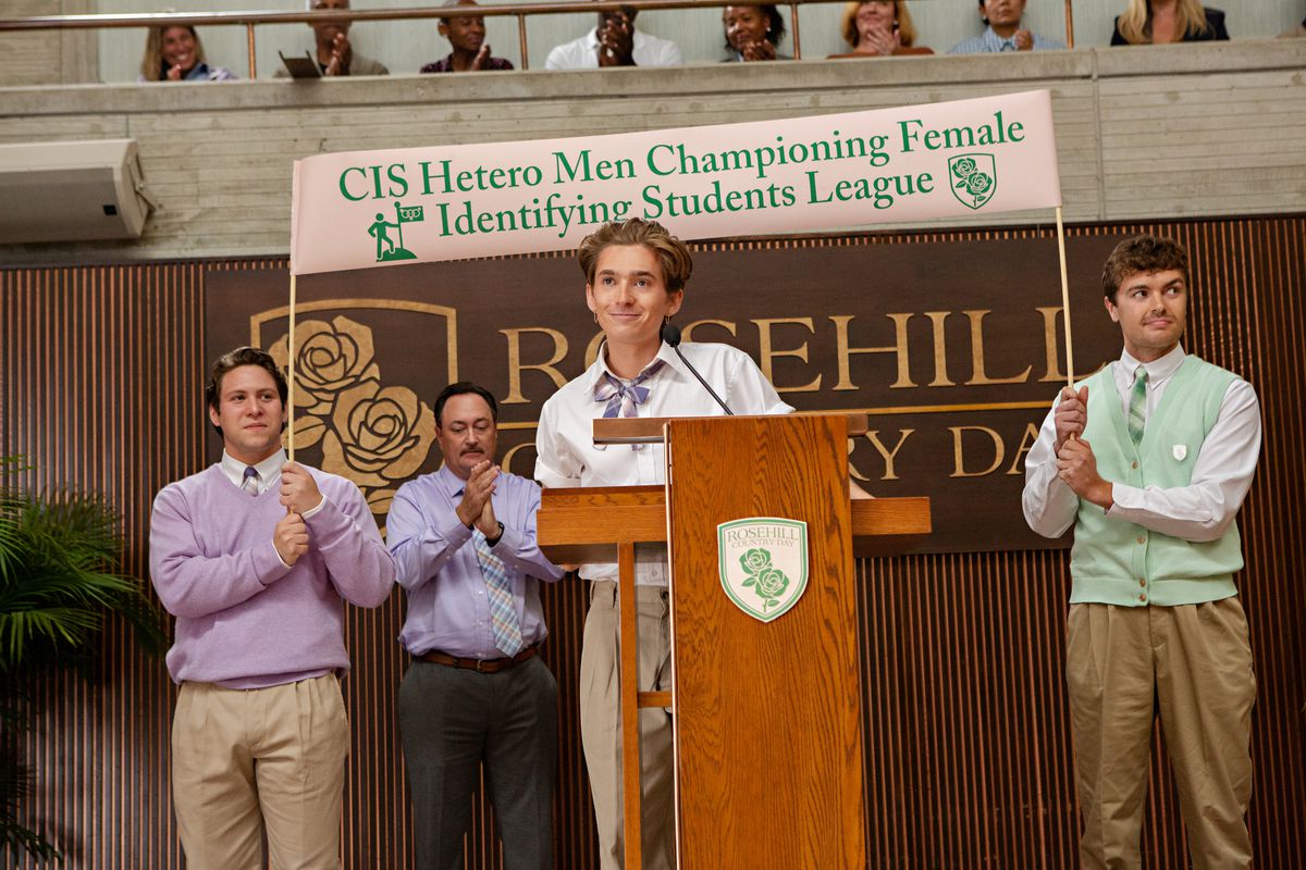 a skinny blonde boy in front of a banner that reads heterosexual men win the schoolgirl identity tournament.  behind him, three men clapped their hands