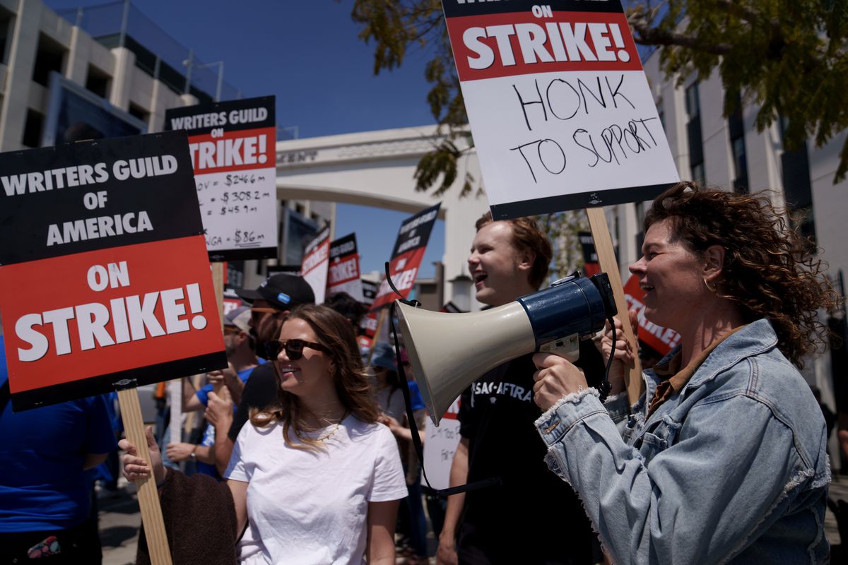 WGA writers striking in front of Sony Pictures Studios, with a woman in the foreground yelling into a megaphone