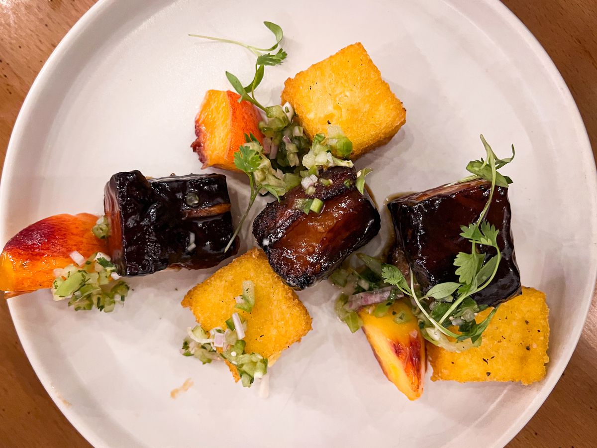 From above, a plate of square slices of glazed pork belly with squares of crispy grits and herbs for garnish.