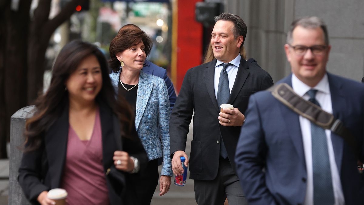 four people dressed in professional business attire are walking on a sidewalk, one is holding a coffee and speaking to a colleague, who is wearing a light blue jacket.