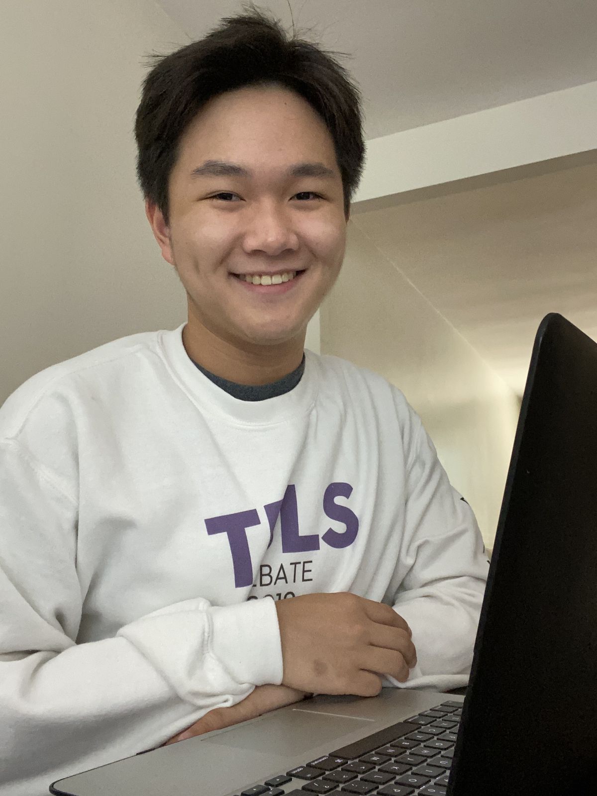 A student wearing a white sweater smiles as he sits at his computer.