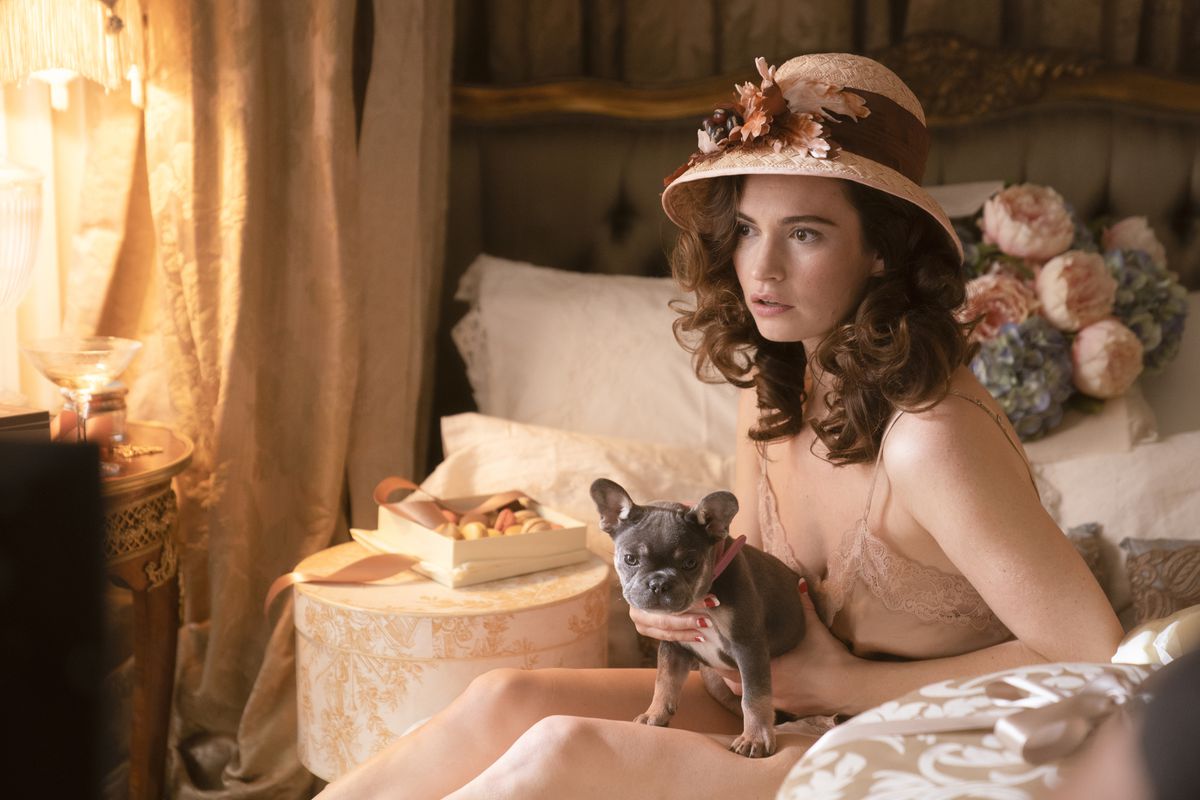 A brunette woman in a lace slip and large cloche hat sits on a bed, holding a lapdog. There’s a box of macarons next to her, and a bouquet of peonies and hydrangeas behind her.