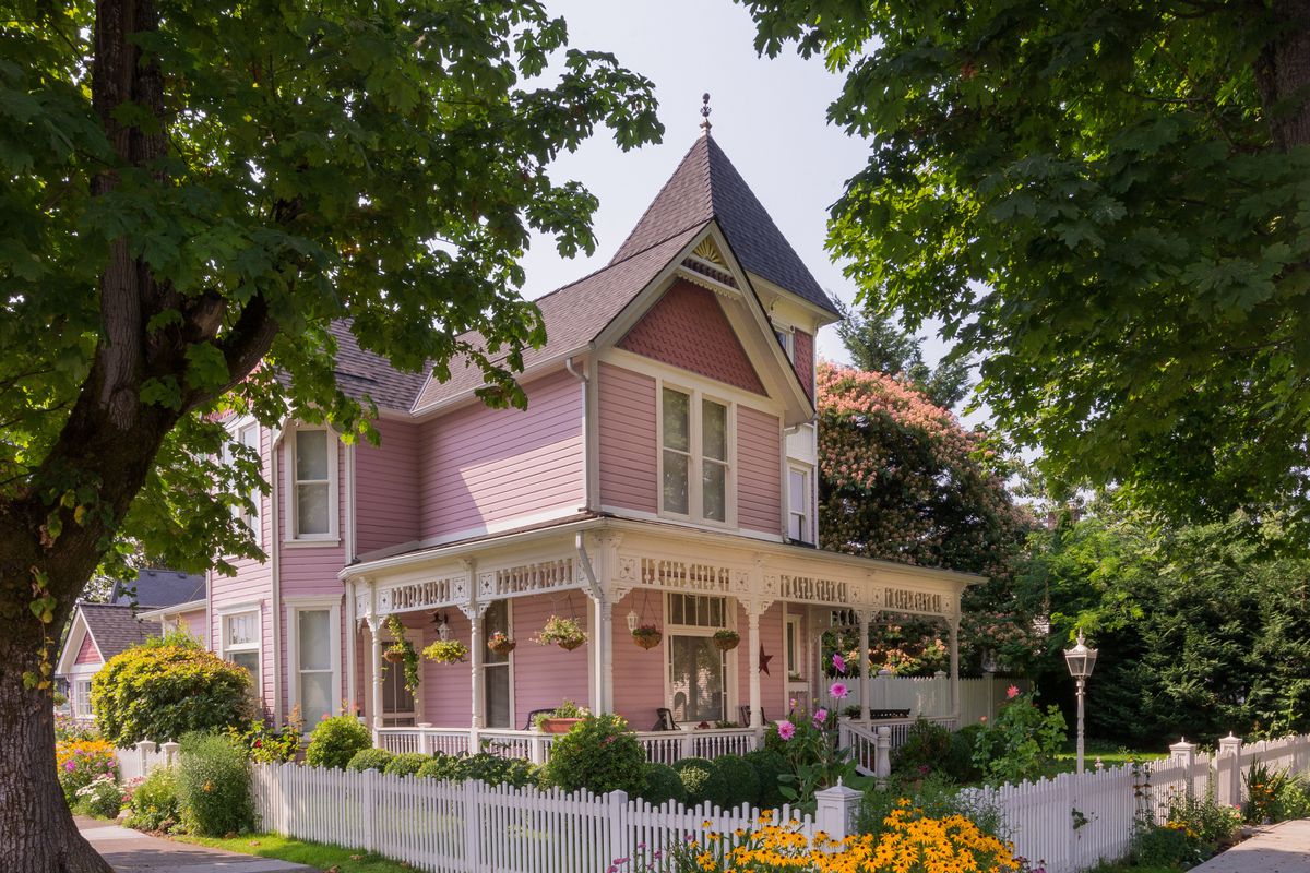 A pink Victorian house with white picket fence and yellow blossoming flowers on the corner lot of a neighborhood with sidewalks and large, green mature trees.