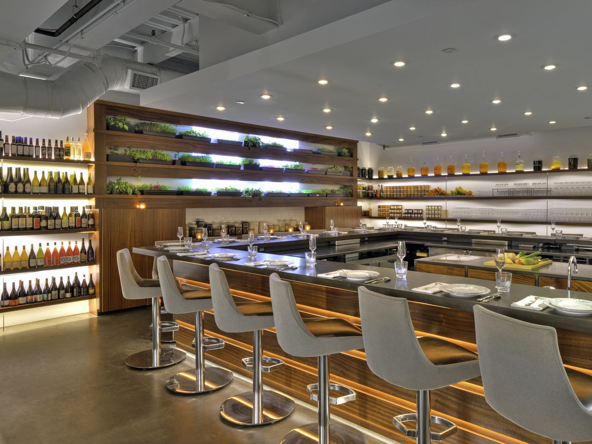 A restaurant interior features a sleek bar, white and light wood accents, shelves of wine, and small planters of herbs.