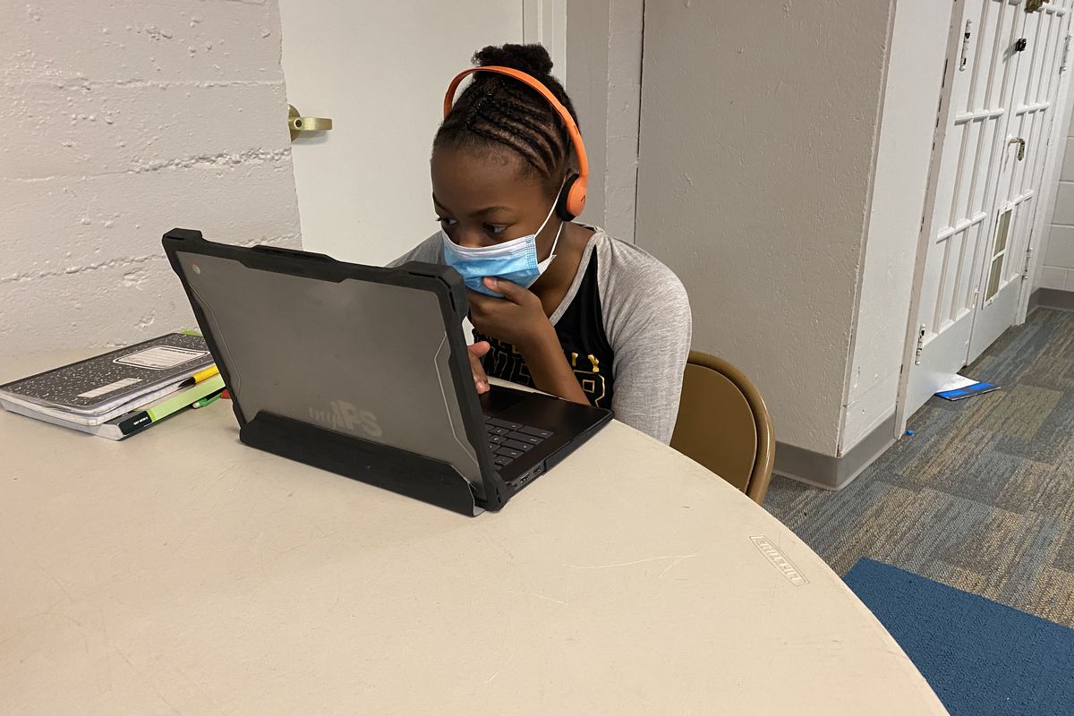 A student with a mask covering her face sits at a table looking at a laptop screen