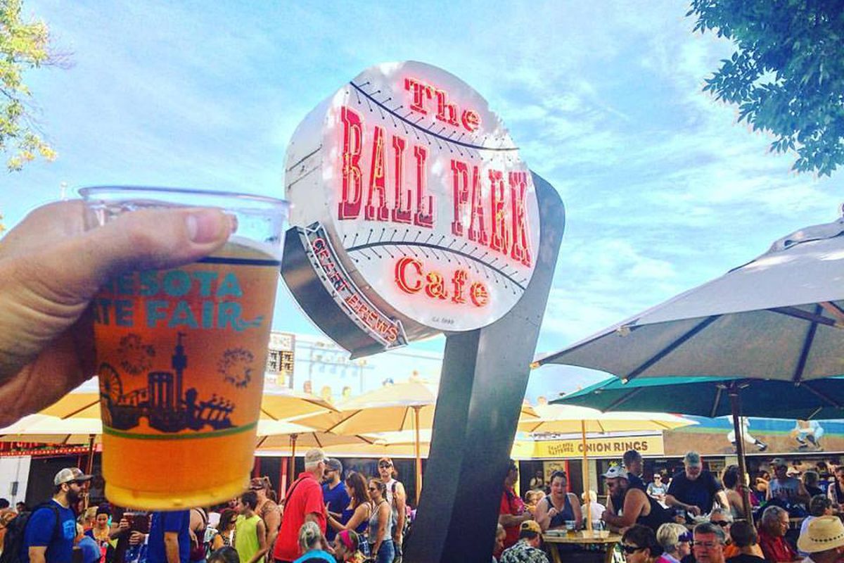A hand holds up a beer next to the baseball shaped neon sign for the Ball Park Cafe on a busy fair day