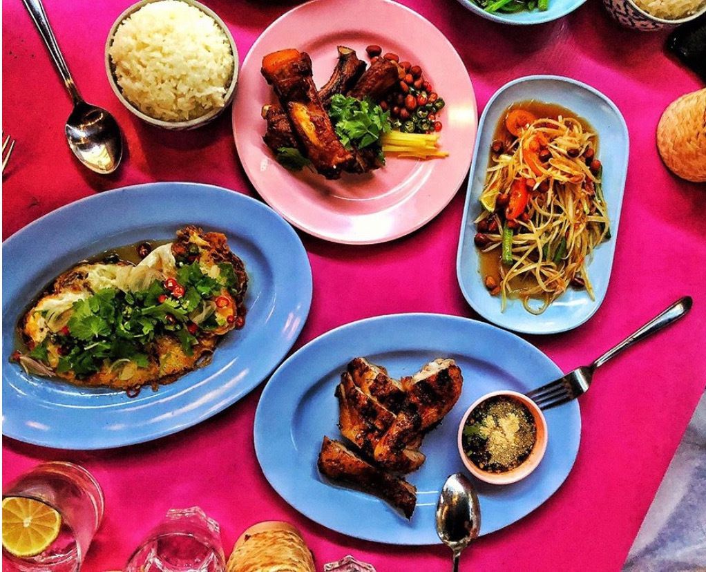 From above, a bright pink table set with various dishes including roast chicken, noodles, ribs, and rice, with condiments and spoons laid out nearby