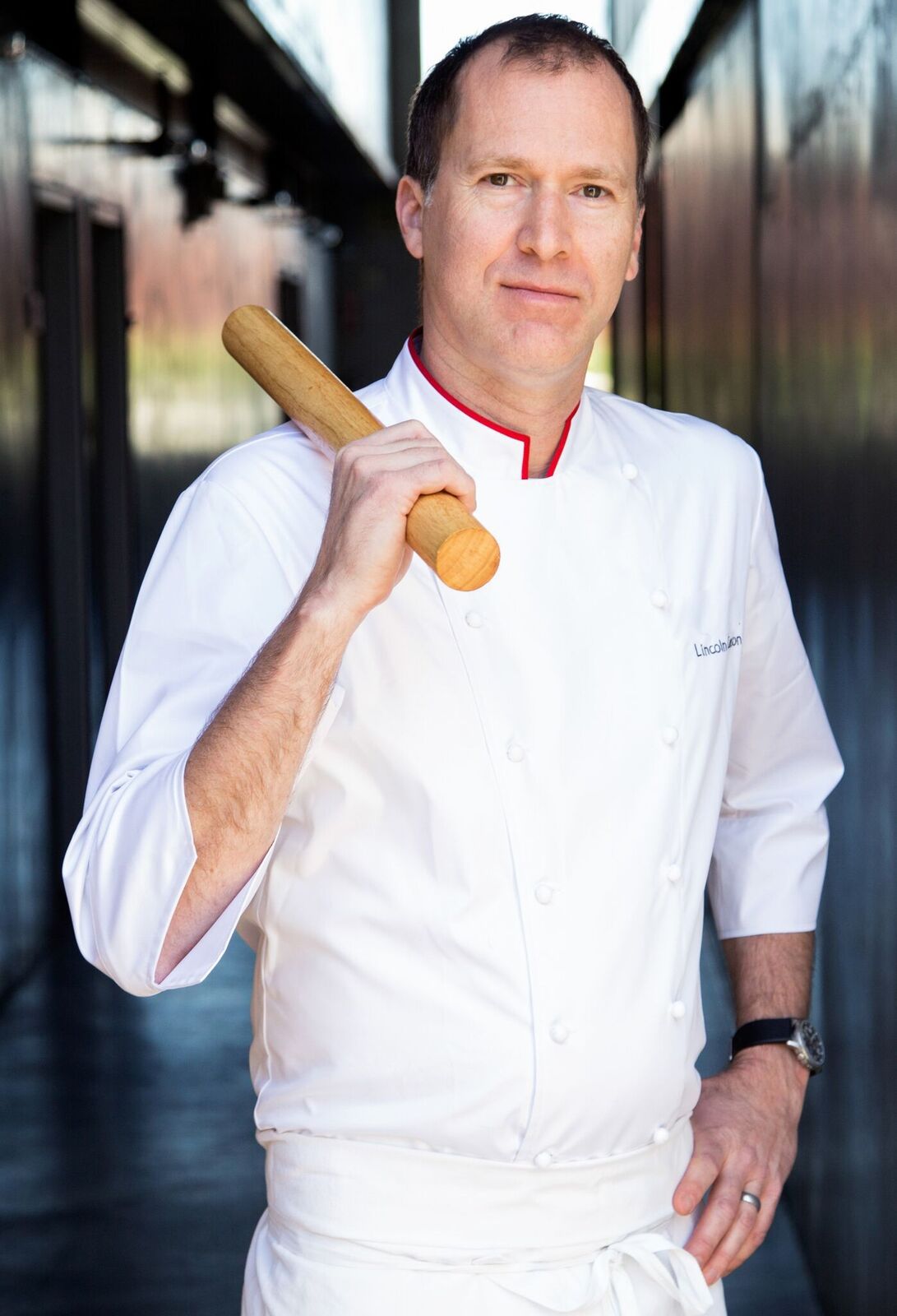 Tall chef Lincoln Carson stands in whites holding a rolling pin.