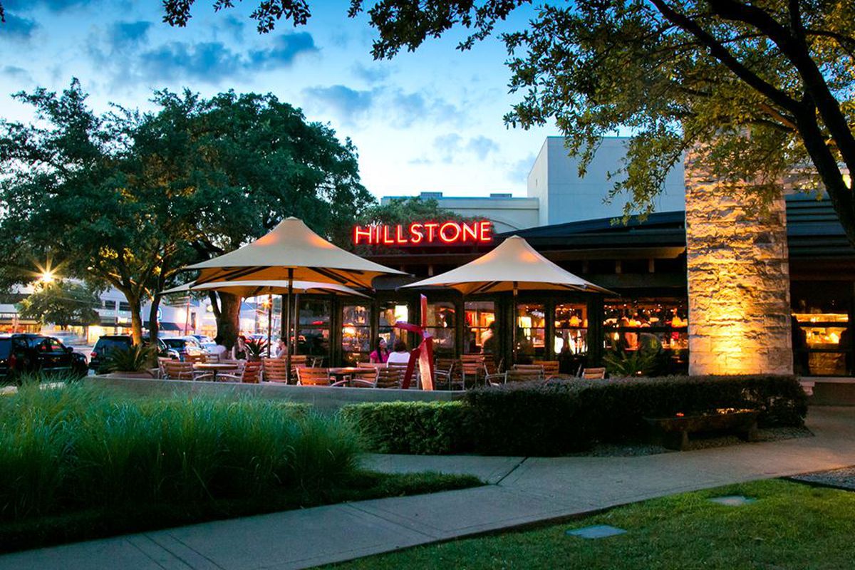 The exterior of Hillstone restaurant at night, with a red and yellow neon sign with its name, a front-lit palm tree in the foreground, and a patio with tan umbrellas. 
