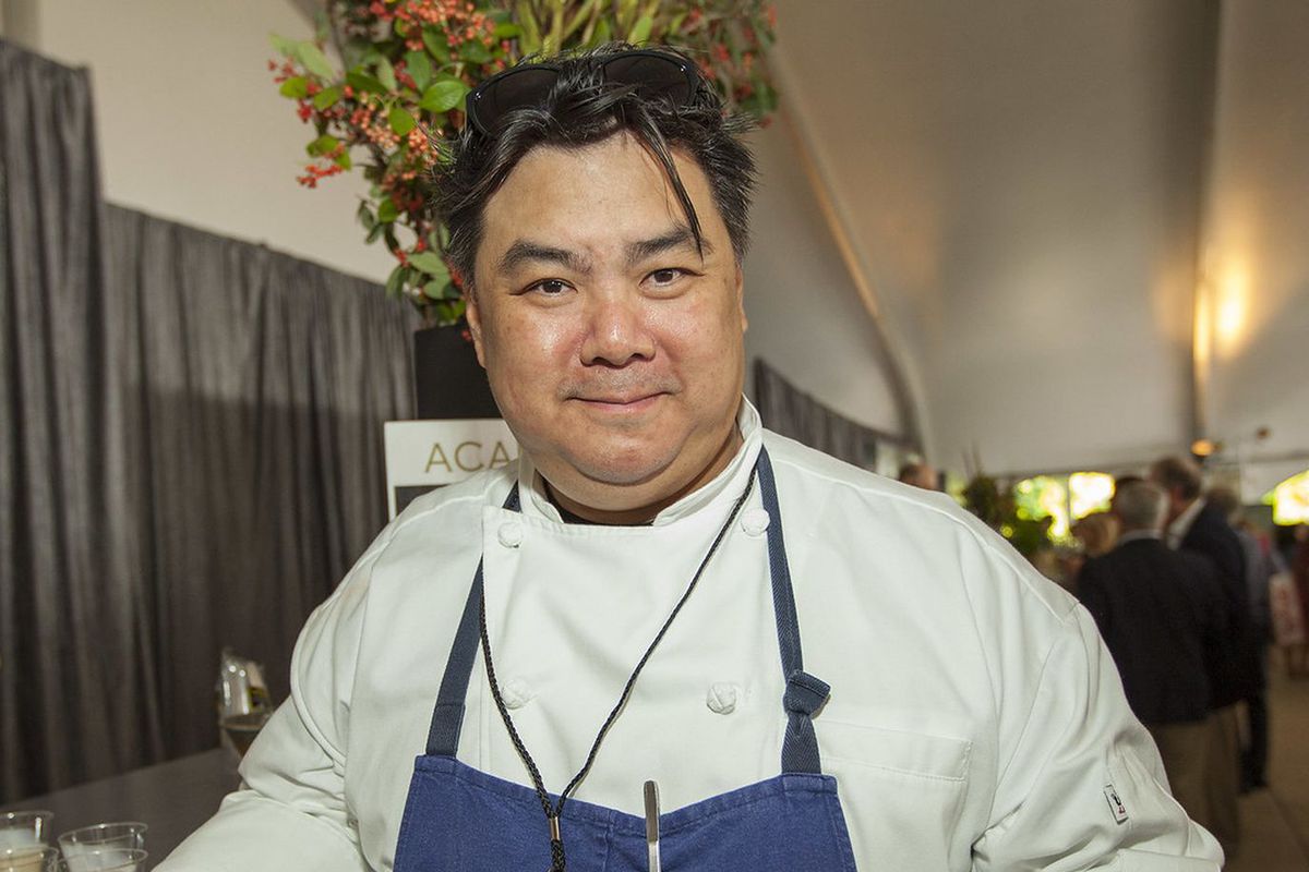 A chef leaning on a table with a drink in his hand wearing a blue apron.