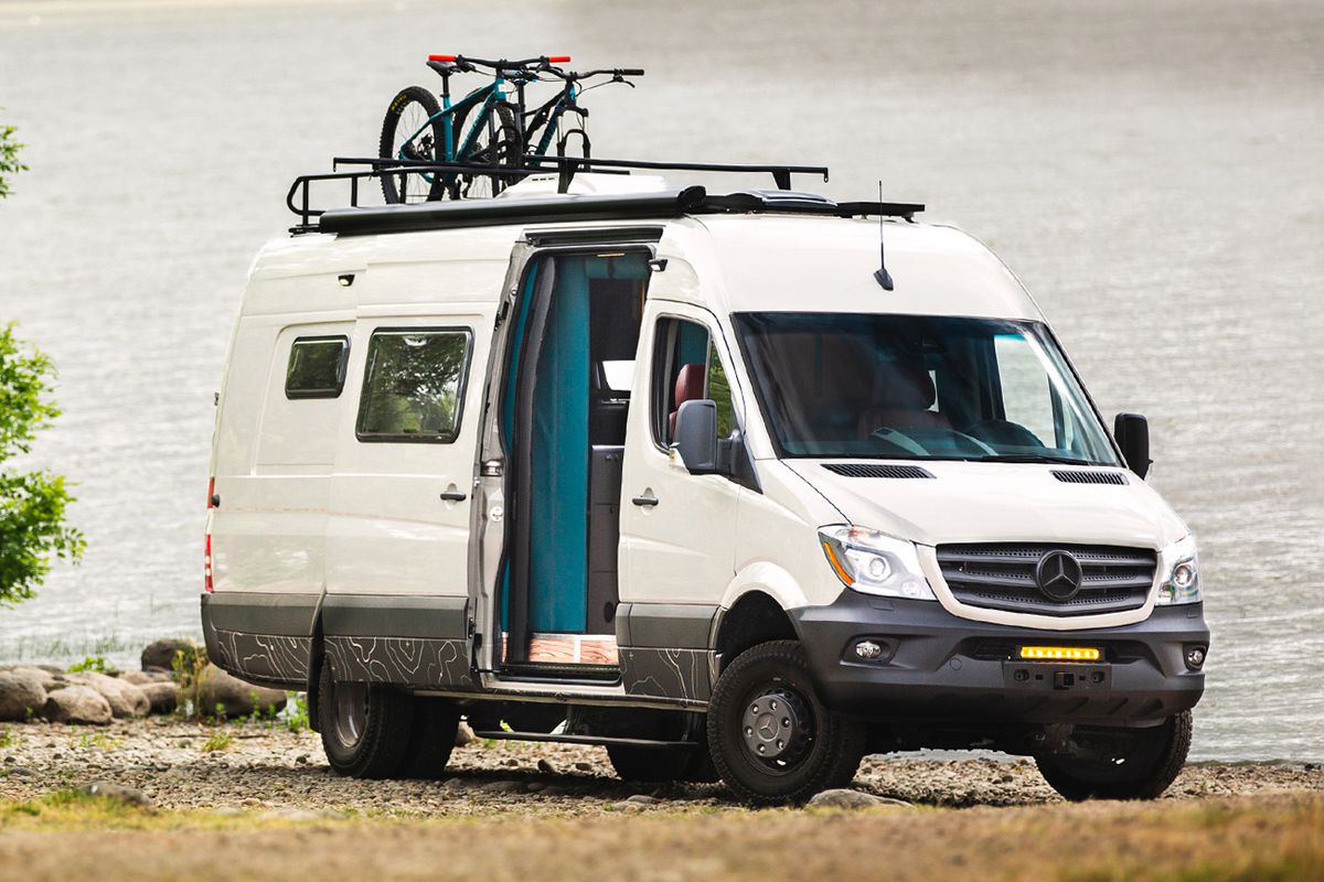 A white camper van sits on the banks of a lake. The camper van’s side door is open and there is a roof rack with two bikes on top. Van life