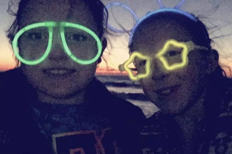 Two young girls, their faces blurry in the dark, wear light-up glow stick glasses as the sun sets behind them.