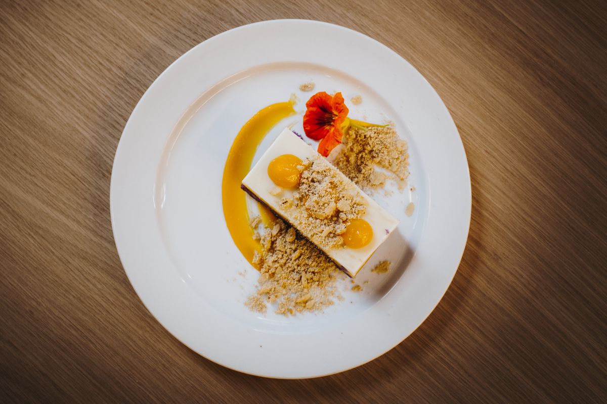 A rectangular cake slice with white frosting and yellow dots of fruit compote covered in crumble with an orange-colored flower on the plate.