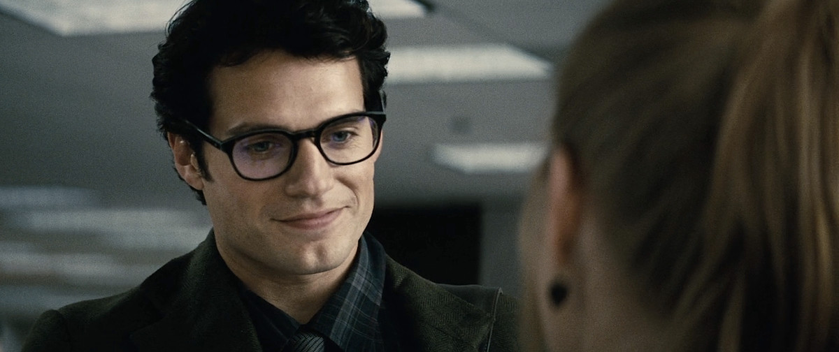 Henry Cavill as Clark Kent stands in the Daily Planet news room and smiles a little shyly and looks down as Lois Lane (Amy Adams) welcomes him to “the Planet” in Man of Steel