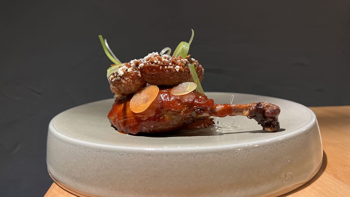 Glazed duck leg gabli sits on a grey concave plate with a sugar-dusted cruller on top
