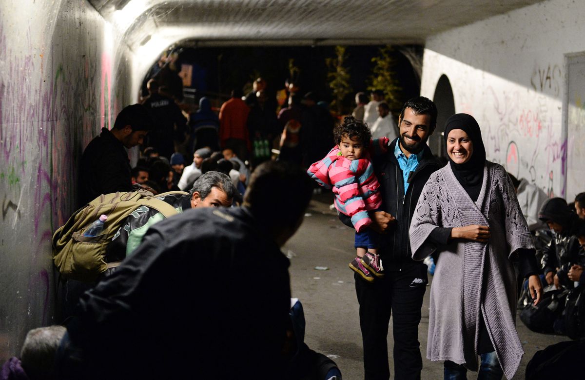 A bearded man in a dark jacket and a bright blue shirt holds a baby in a puffy pink and blue striped coat; a woman in a black hijab and grey sweater walks next to him. Both the man and woman are smiling. They pass groups of refugees, sitting in the dim light of a white walled shelter covered in graffiti.  