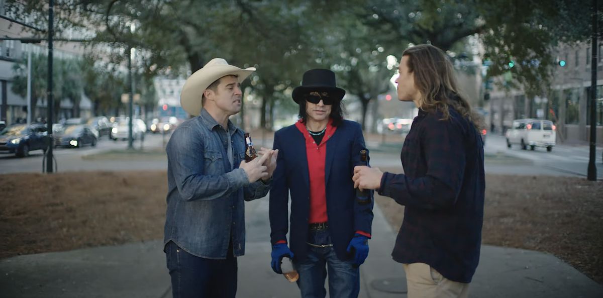 Patrick (Tommy Wiseau, in flat-top fedora, huge sunglasses, a bright red shirt, and inexplicable blue gloves) stands between his buddies Tim (Isaiah LaBorde, in jean jacket and a cowboy hat), and Georgie (Mark Valeriano, in khakis and a button-down shirt) on a New Orleans sidewalk as they argue about the shark in Wiseau’s movie Big Shark