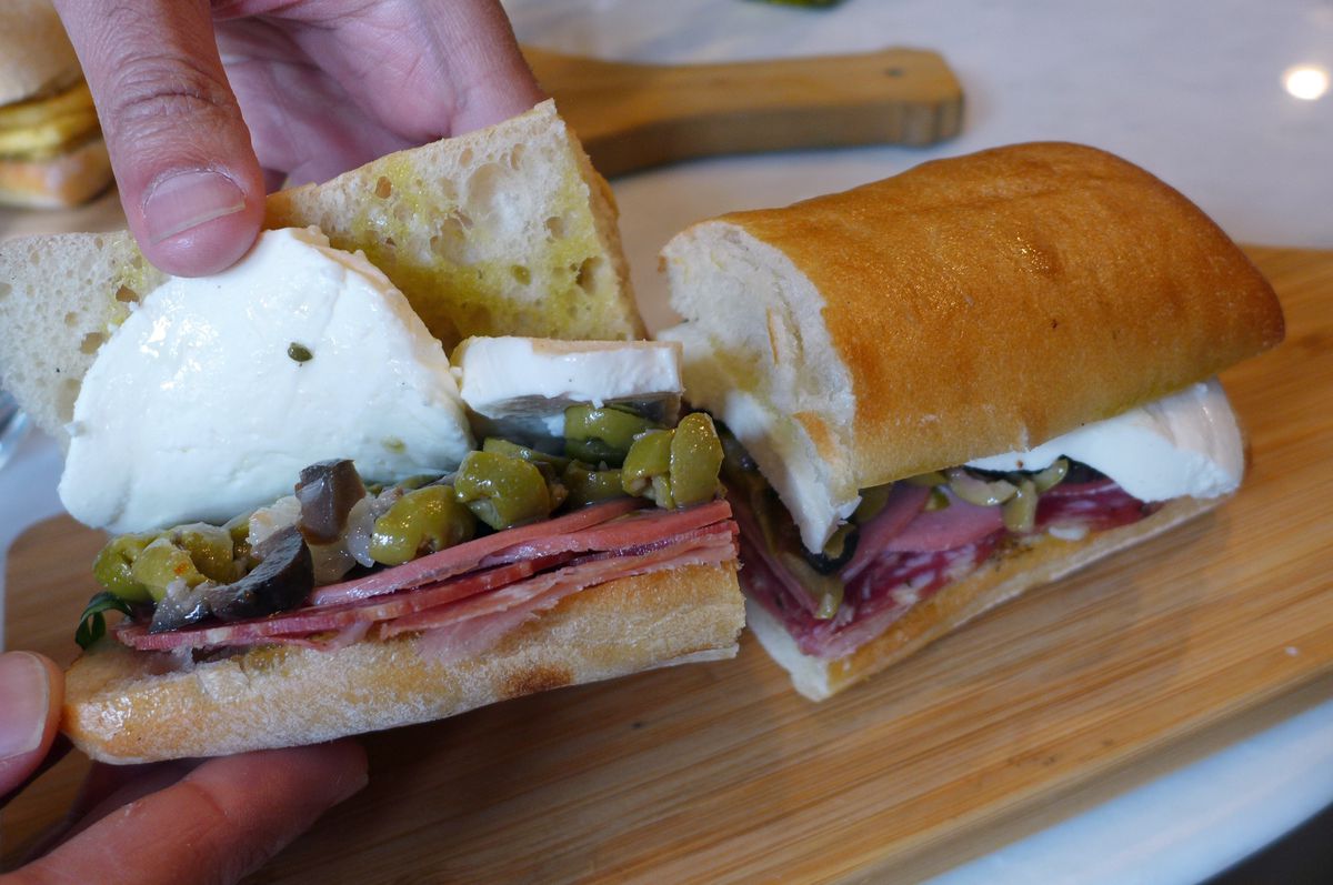 A hero with cold cuts, rounds of bright white mozzarella, and green olives with one half held open by a gnarled hand.
