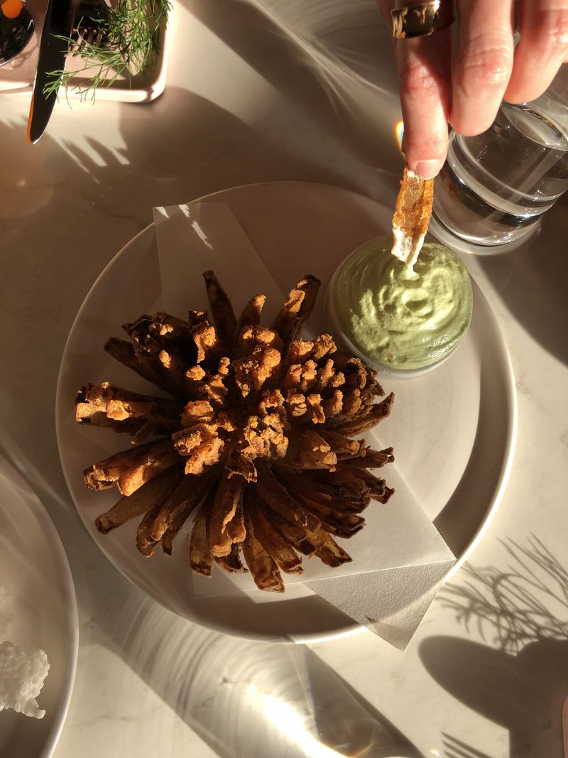 An overhead view of a large, blooming fried onion, with a hand dipping a piece into coconut dip.