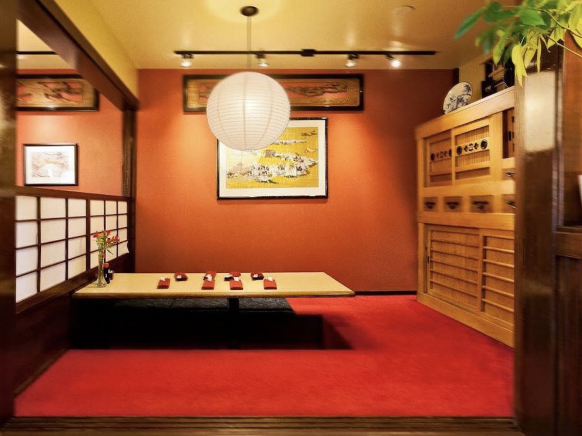 A low chabudai-style table in a private dining area partitioned on one side by a paper screen, lit by a large paper lantern above, with a Japanese print on the back wall, and a chest to the other side