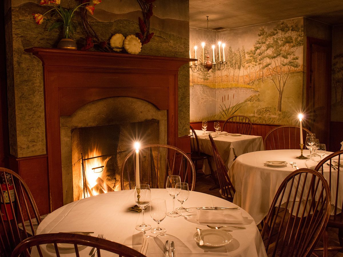 A dining room with a fire in the fireplace, idyllic wallpaper, dark wood paneling, tables with rustic chairs set for dinner with candles