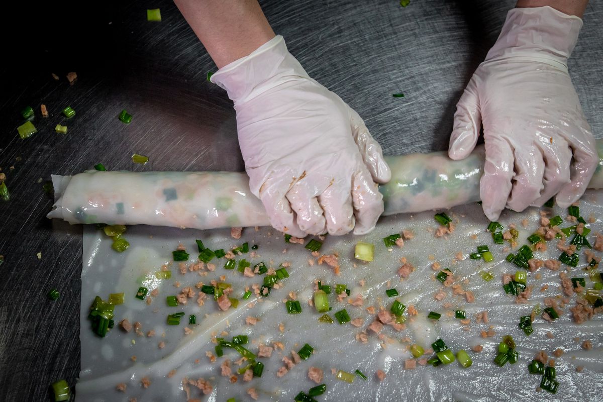 Gloved hands roll rice rolls, which are topped with pieces of pork and scallions.