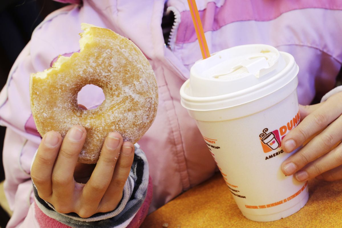 A person holding a donut with a bite taken out of it in one hand, and a styrofoam cup of dunkin donuts coffee in the other