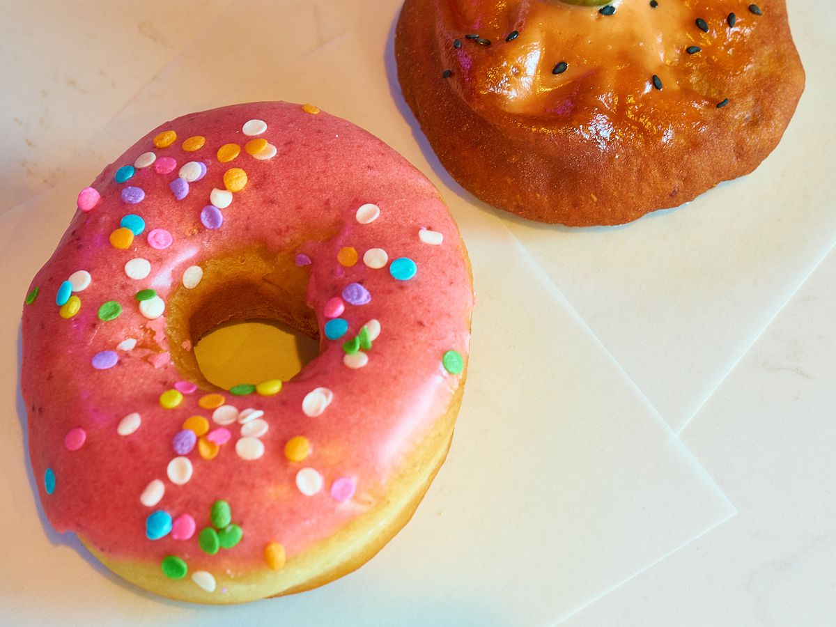 A bright pink vegan donut with sprinkles, next to a brown hole-less donut topped with a pickle.