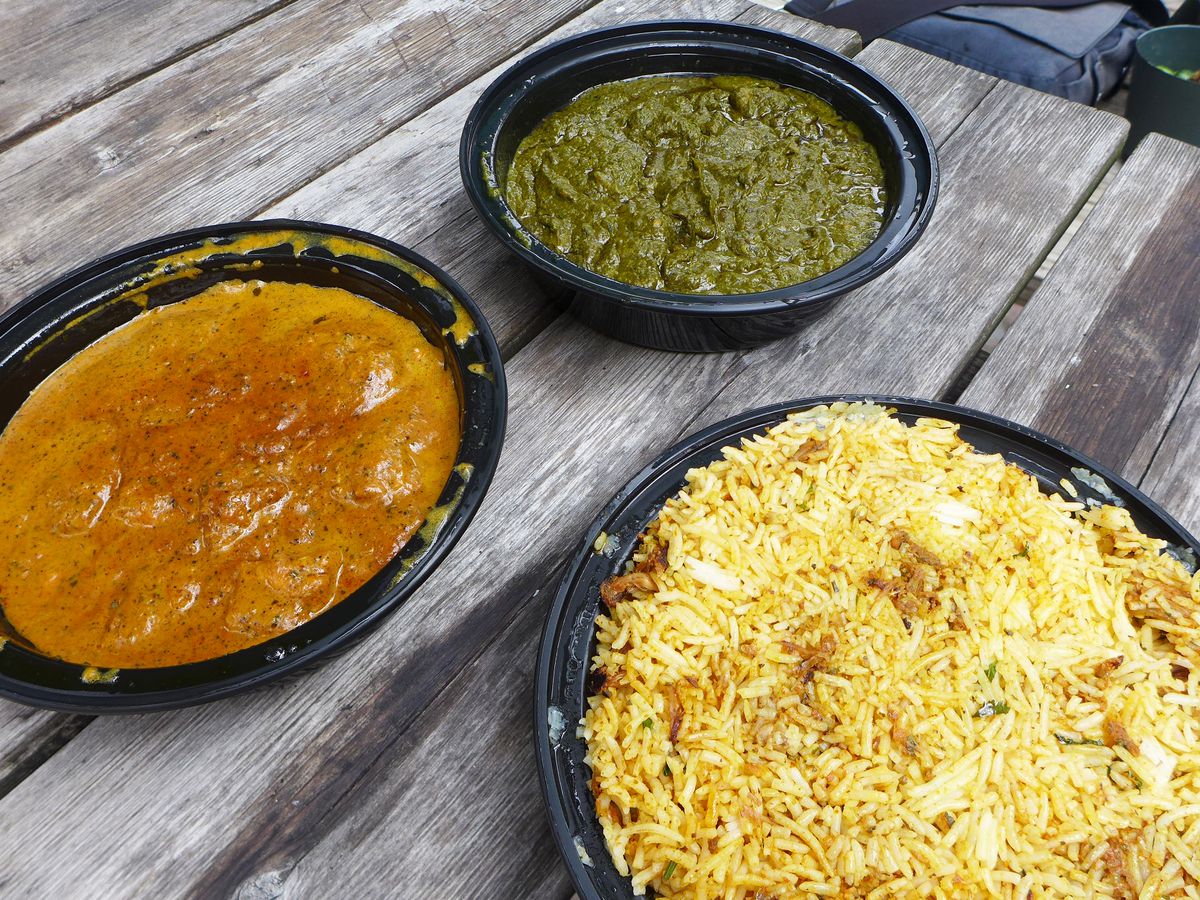 Three Indian dishes in plastic containers on a worn picnic table top, one green, one brown, and one rice based.