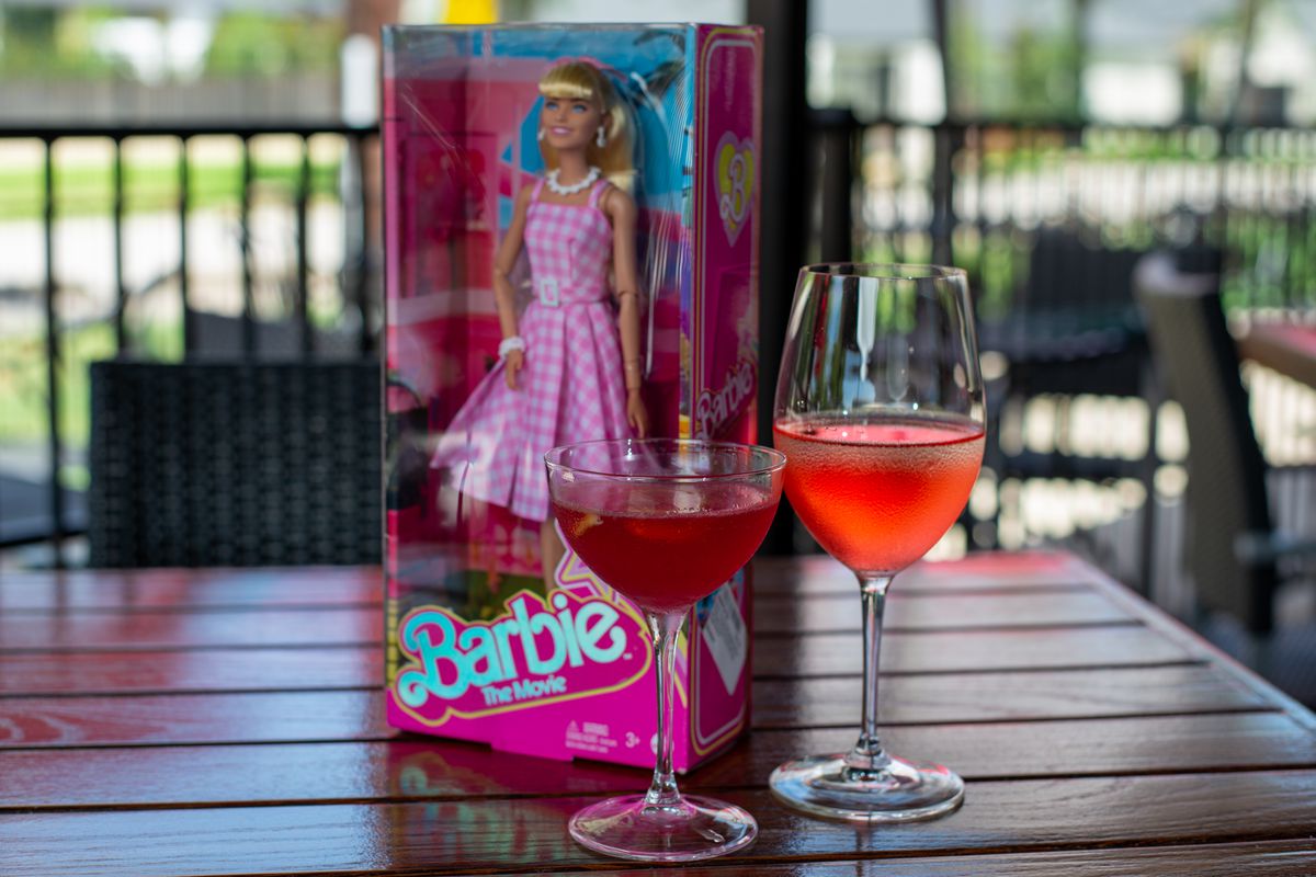 A Barbie doll in a box sits on the table with two glasses of wine.