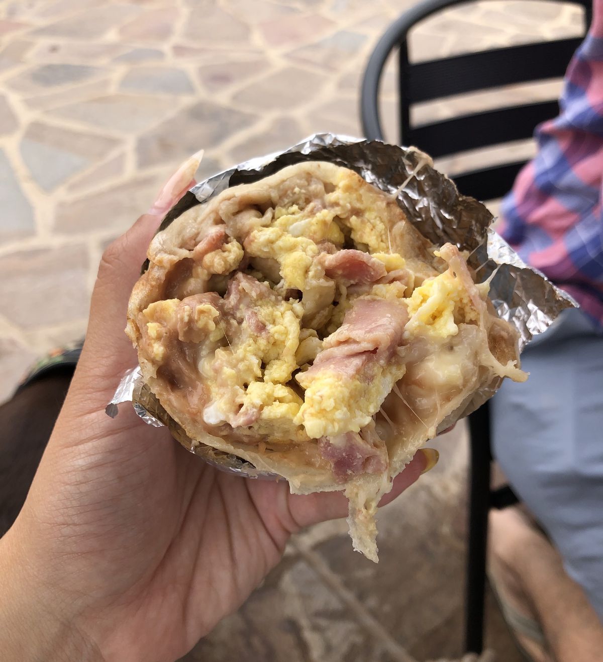 A hand holding a photo of half a breakfast burrito encased in tinfoil with slices of ham, refried beans, scrambled egg, and melted cheese.