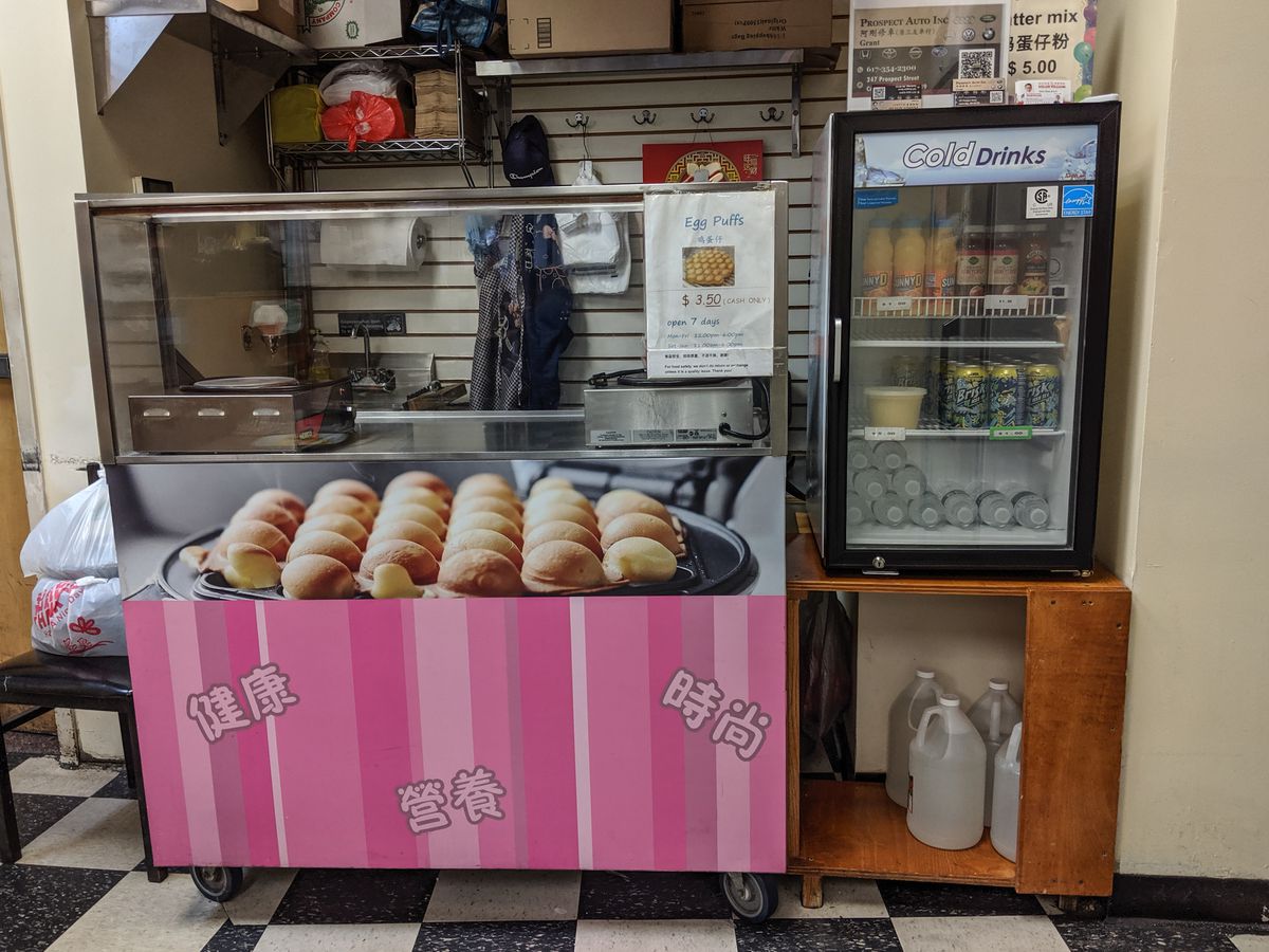 A pink cart that serves egg puffs inside of a small food court. It’s decorated with a large photo of egg puffs.