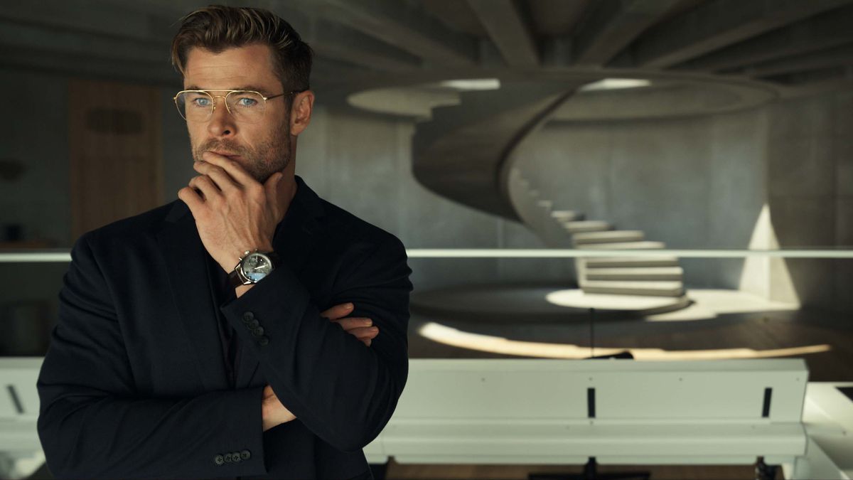 Chris Hemsworth ponders musingly, or muses ponderously, in the foreground of a barren concrete room with an elegant white spiral staircase in the background. Also an out-of-focus Miles Teller.