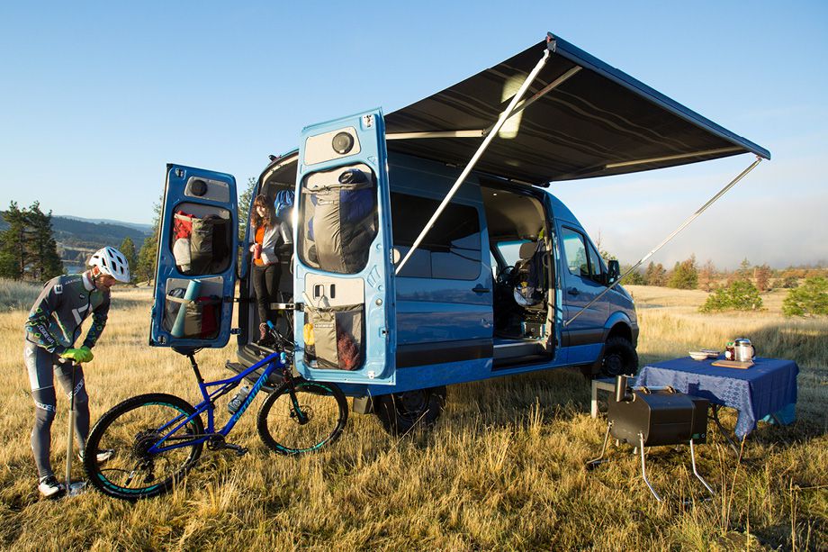 A blue camper van. The doors are open and there is an attached awning. There is a man pumping air into a bicycle near the van. A woman stands in the back of the van. There is a table and a portable grill under the awning. 