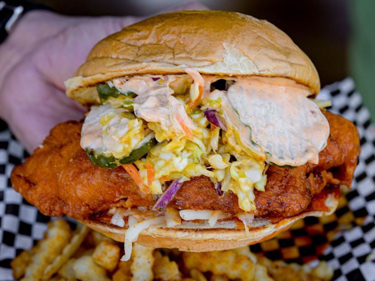 A white hand holds a fried chicken sandwich overflowing with pickles and coleslaw on a brioche bun above a basket with checkered paper and seasoned crinkle cut fries.