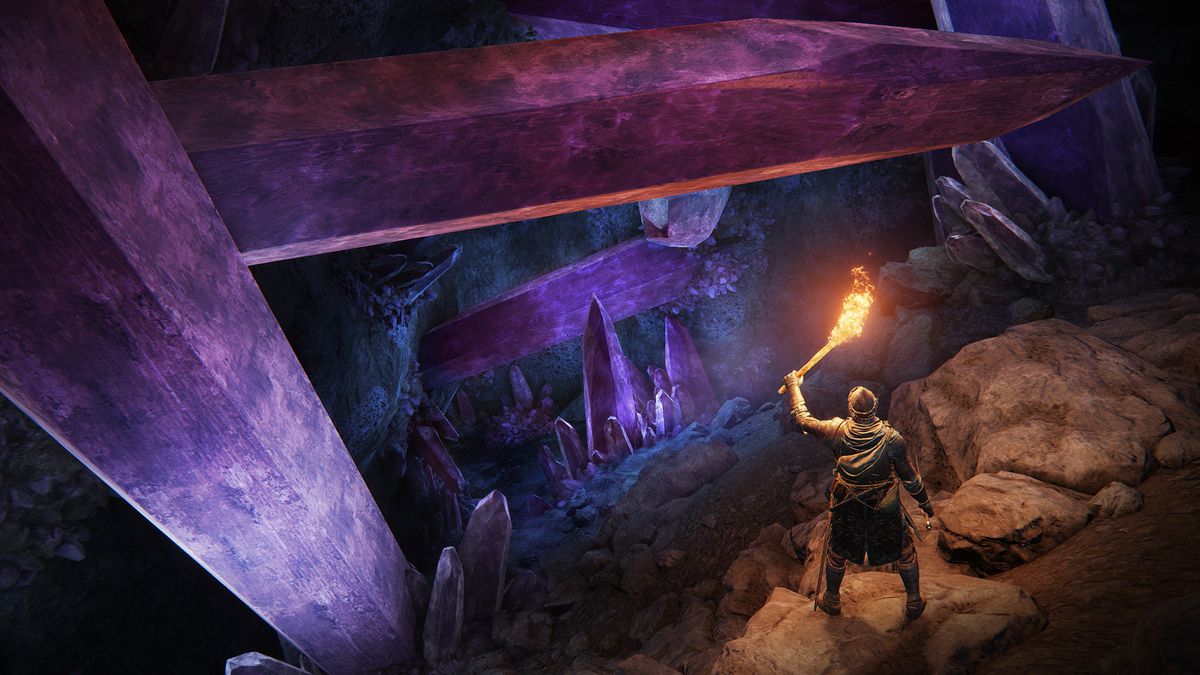 A Tarnished holds a torch aloft and looks down at a crystal cave in a screenshot from Elden Ring