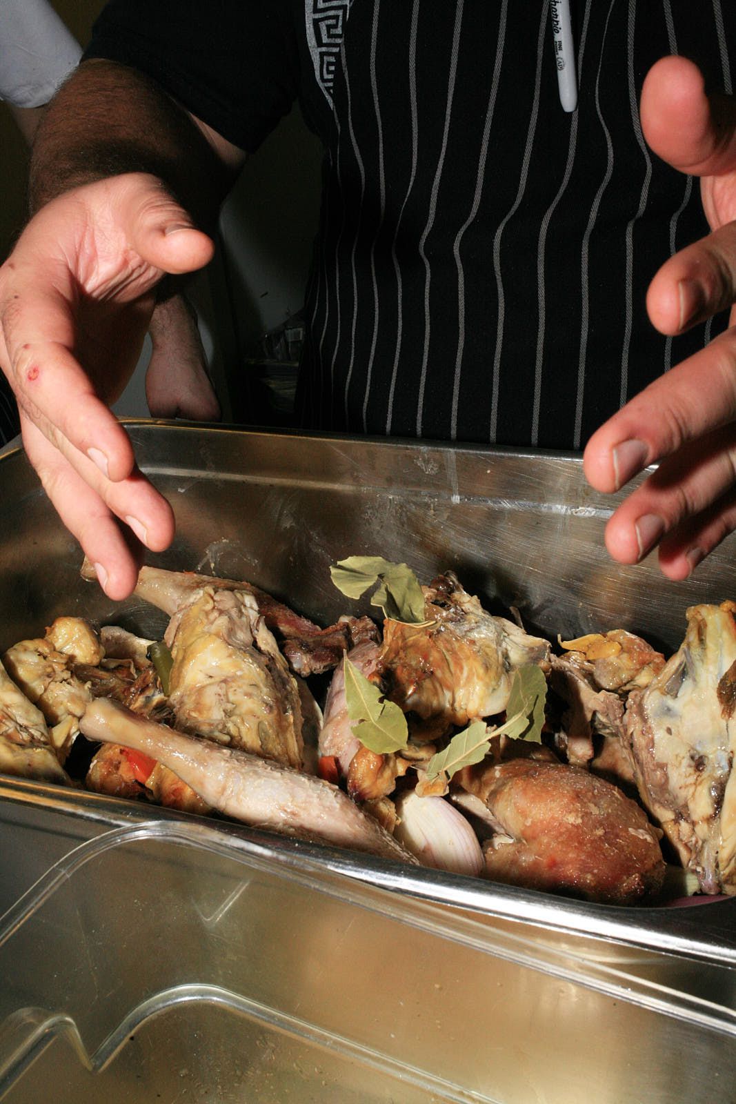 A closeup of a chef’s hands dropping bay leaves into a metal tray of roast duck legs and vegetables.
