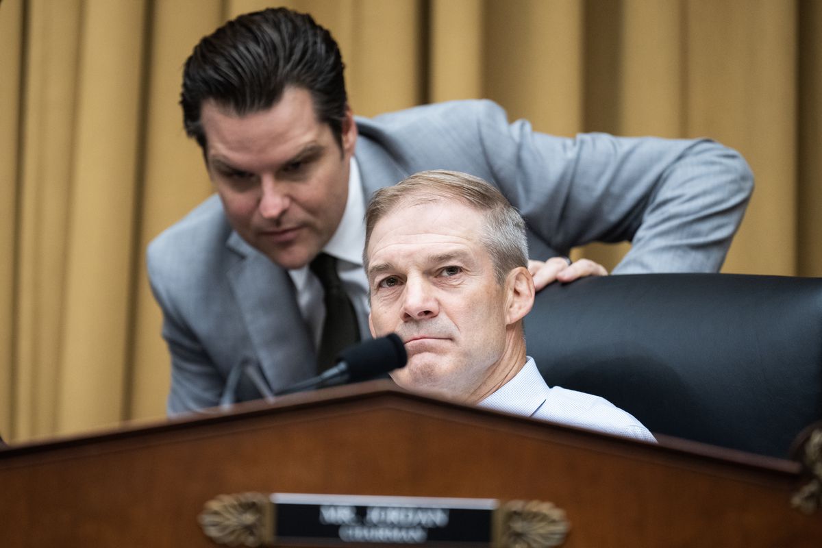 Matt Gaetz, in a light gray suit, leans over the back of Jim Jordan’s chair and talks to him, in a congressional meeting room.