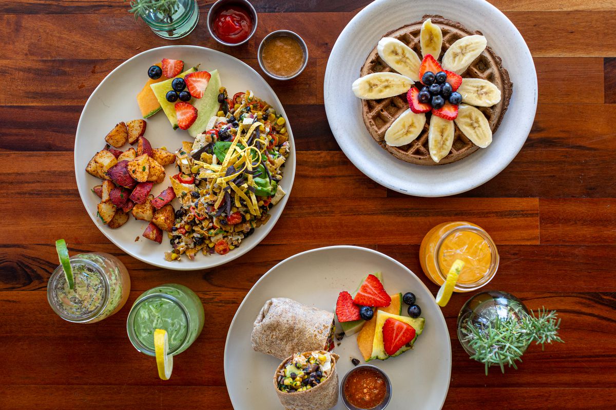 An overhead shot of colorful drinks, a burrito, a breakfast plate, and a waffle topped with banana slices.