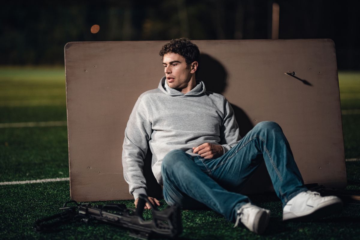 Theo James, sitting on a sports field behind an overturned table, surrounded with guns, in a clone battle to the death in Dual